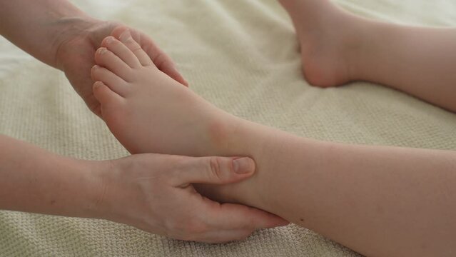 Closeup of unrecognizable doctor doing gymnastics and foot massage to five year old boy. Close up hands of masseuse doing orthopedic leg exercises to little child. Shooting in slow motion.