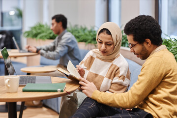Portrait of modern Middle Eastern woman wearing head cover in office and looking at notes with male colleague