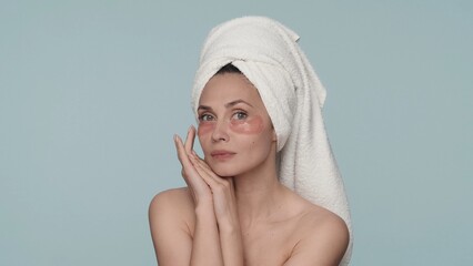 Portrait of a woman with a towel on her head and red hydrogel patches under her eyes close up. A woman enjoys the purity and smoothness of her skin in the studio on a blue background.