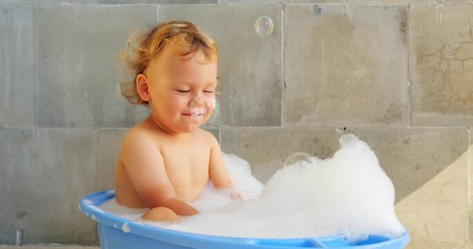 Handsome little boy take bath, sitting in warm water at baby bathtub, slow motion shot. Heap of white fluffy foam on water, large soap bubbles soar down. Short portrait clip of happy child