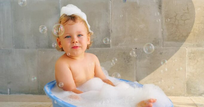 Delightful baby boy sits in warm, soapy baby bathtub, looking mischievously at camera with his tongue sticking out. Slow motion footage captures large soap bubbles flying in foreground