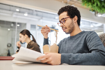 Side view portrait of young Middle Eastern man eating takeout food at office workplace and reading documents, copy space