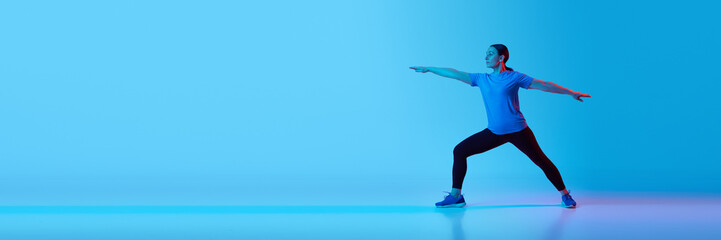 Dynamic postural. Mature woman in black bodysuit training, stretching over blue background in neon light. Concept of sport, healthy lifestyle, fitness, body care, wellness. Banner. Copy space for ad