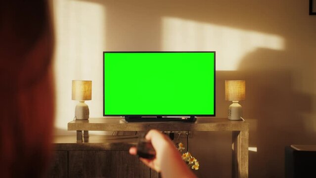 Woman watching TV with green screen in living room. Female person switching channels, using remote control, sitting on sofa.