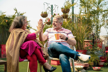 Happy indian senior couple drinking tea or coffee in colorful mugs sitting at garden.