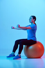 Smiling mature woman sitting on fitness ball and training with dumbbells against blue studio background in neon light. Concept of sport, healthy lifestyle, fitness, body care, wellness, ad
