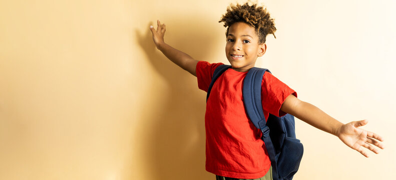 Back To School! An African Boy With Afro Hair Poses On A Brown Studio Background In Casual Clothes And A Backpack. The Child Between 5 And 6 Years Old Has An Expression With Open Arms.