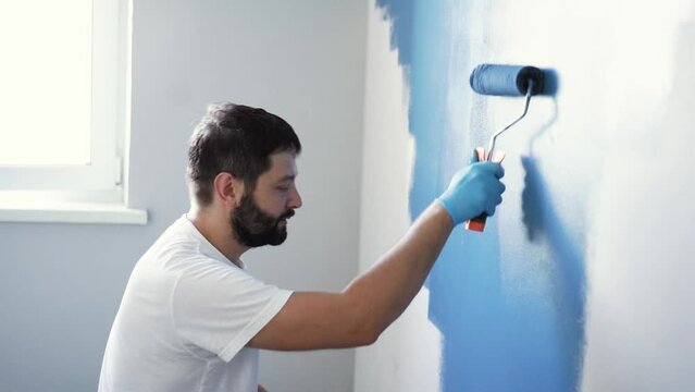caucasian bearded man painting wall with paint roller. Painting apartment, renovating home with blue color paint. High quality FullHD footage