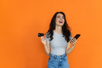 Cheerful woman posing isolated on orange wall background using mobile phone, credit bank card