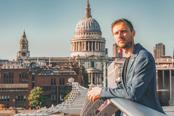 Young man leaning on railing of Millennium bridge. Male tourist looking away during vacation. Cathedral of St. Paul Church with clear blue sky in background on sunny day. - obrazy, fototapety, plakaty