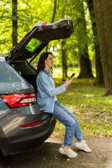 Young happy female sitting in car trunk and looking away while having phone conversation