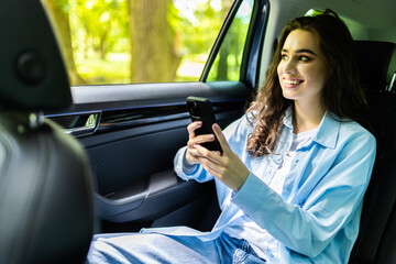 Beautiful woman is using a smart phone and smiling while sitting on back seat in the car