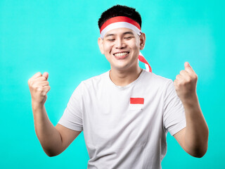 Naklejka premium A portrait of an Asian man celebrating Independence Day, wearing a white t-shirt and a red and white headband, showing a victory pose with his hands raised up. Isolated with a turquoise background.