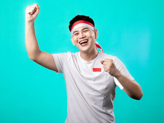 A portrait of an Asian man celebrating Independence Day, wearing a white t-shirt and a red and white headband, showing a victory pose with his hands raised up. Isolated with a turquoise background.