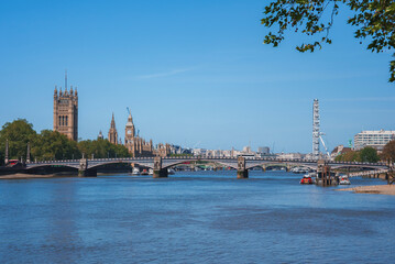 Big Ben and Millennium Wheel at banks of river. Westminster bridge over Thames water with blue sky in background. Scenic view of famous tourist attraction in city during sunny day.