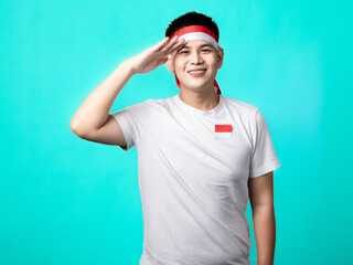 Fototapeta premium A portrait of an Indonesian (Asian) man celebrating Independence Day, wearing a white t-shirt and a red and white headband, in a salute pose. Isolated with a turquoise background.