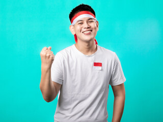 Fototapeta premium A portrait of an Asian man celebrating Independence Day, wearing a white t-shirt and a red and white headband, showing a victory pose with his hands raised up. Isolated with a turquoise background.