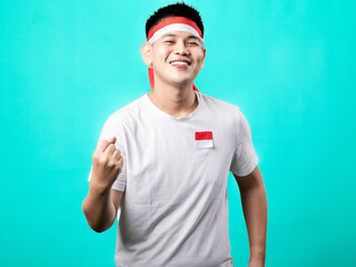 Fototapeta premium A portrait of an Asian man celebrating Independence Day, wearing a white t-shirt and a red and white headband, showing a victory pose with his hands raised up. Isolated with a turquoise background.
