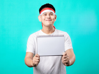 Portrait of an Indonesian Asian man celebrating Independence Day, wearing a white shirt and a red-and-white headband, seen in a pose with an empty paper, isolated with a turquoise background.