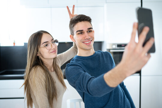 Smiling teenage girl and boy taking a selfie with smartphone. Girl holding two fingers behind his boyfriends head. April fool day holiday concept
