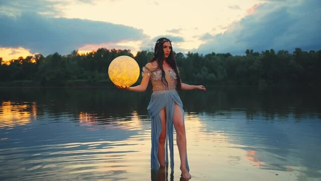 Fantasy woman holding moon magic light glowing planet in hand. Girl goddess night elf beauty face blacl long hair, river nymph wet blue dress, stands on water dark sky clouds lake. Lady fairy art. 4k