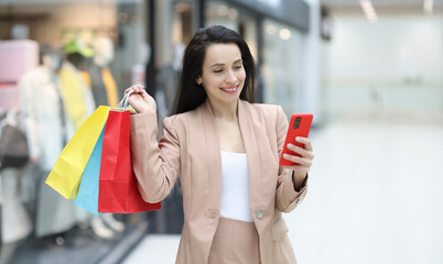Smiling woman holds in her hand smartphone and purchases in mall. Retail therapy and shopping mood concept