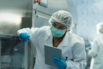 Naklejka premium Female worker wearing protective mask inspecting quality of plastic water tank on conveyor belt in drinking water factory. Woman worker using tablet checking bottle or gallon on conveyor