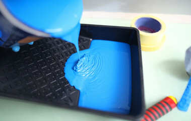 Hand pour blue paint from can into black plastic tray. In background there are two masking tape of different width.