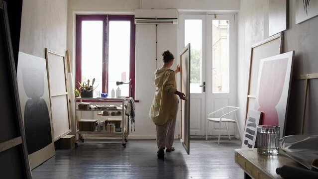 A young artist arranges her paintings in her studio. The woman carries a large canvas and places it against the wall. Workshop of a contemporary artist, abstract painting, art.