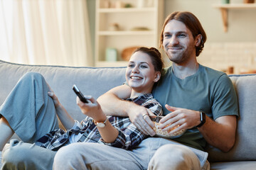 Portrait of smiling young couple watching TV together sitting on sofa and holding remote control