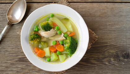 Chicken soup with broccoli, green peas, carrots and celery in a white bowl on a wooden background in rustic style. Top view