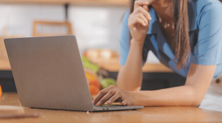 A young woman with a beautiful face in a blue shirt with long hair eating fruit sitting inside the kitchen at home with a laptop and notebook for relaxation, Concept Vacation.