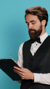 Vertical video Bellboy reading checklist of guests, holding ledger or register on clipboard to verify list of travellers arriving at hotel. Staff checking reservations in official catalogue to keep