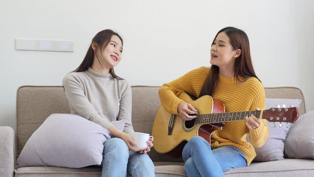 Activities of LGBTQ Asian female lover couple sat on the couch smile happily sing a song and play guitar dancing to the music at living room. Lesbian, Transgender. LGBTQ concept