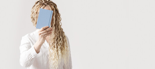 Woman holding blue covered paper book in front of face, light background, no face trend concept....