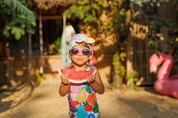 Funny cute girl on summer vacation. The child has fun on the beach. Cute baby girl in a colorful swimsuit and sunglasses is resting and eating fruit.