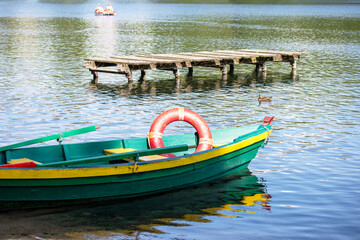 
A green-yellow wooden fishing boat with a red life ring on a background of water and a wooden boardwalk