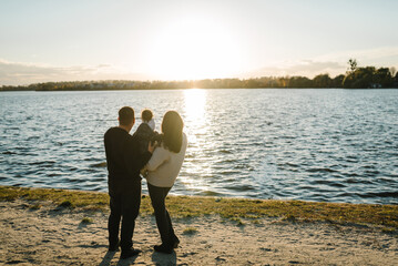 Family spending time together on beach at sunset on vacation. Mom, dad hugs child, walking in park. Concept of autumn holiday. Mother, father and daughter near lake in nature. Family photo. Back view.