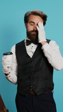 Vertical video Doorman holding coffee drink in studio, wearing white gloves and feeling confident working in hotel industry. Bellhop worker in suit and tie drinking beverage next to trolley bags.