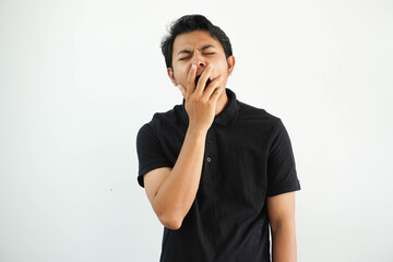 sleepy young asian man yawning by covering mouth with hand wearing black polo t shirt isolated on white background