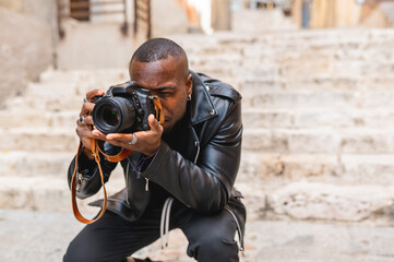 Concentrated African American male photographer taking pictures on professional photo camera while sitting on haunches on old city stairs