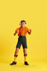 Full-length of little boy, child in stylish clothes listening to music in headphones against yellow studio background. Concept of childhood, kids emotions, fashion, hobby, ad