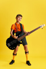 Obraz premium Boy, child in stylish clothes playing guitar against yellow studio background. Music and education. Concept of childhood, kids emotions, fashion, hobby, fashion and lifestyle, ad