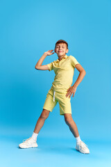 Full-length of cheerful little boy, child in casual clothes posing, dancing against blue studio background. Relaxation. Concept of childhood, kids emotions, holiday, summer, fun and joy, ad