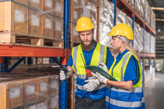 male employee carries a scanner to count boxes on a shelf, and a female employee checks inventory, both wearing reflective uniforms and helmets, walking through a distribution center.