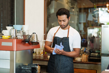 Asian man with beard on face, wearing apron, standing, using cloth clean glass absorb water from washing cleaning, prepare open cafe, make coffee for customers come cafe drink coffee in morning...