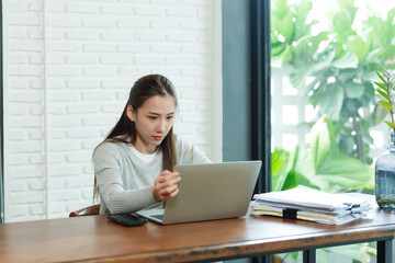 Asian woman is working in favorite cafe with cup coffee, where always takes job, sat down open pile documents on table, bent down type on laptop that lay, deliberately with work laid out in front..