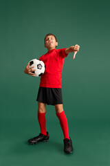 Fototapeta na wymiar Full-length image of little boy, child in red uniform posing with football ball against green studio background. Concept of childhood, kids emotions, sportive lifestyle, action, hobby, ad