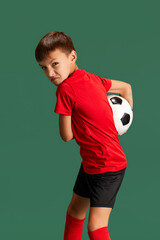 Fototapeta na wymiar Future football player. Little boy, child in sportswear posing, playing with ball against green studio background. Concept of childhood, kids emotions, sportive lifestyle, action, hobby, ad