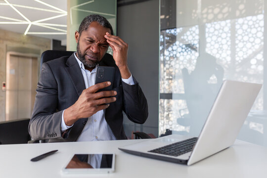 Upset and disappointed man inside office holding phone, businessman reading bad news at workplace inside office, african american senior boss unhappy with achievement results notification.